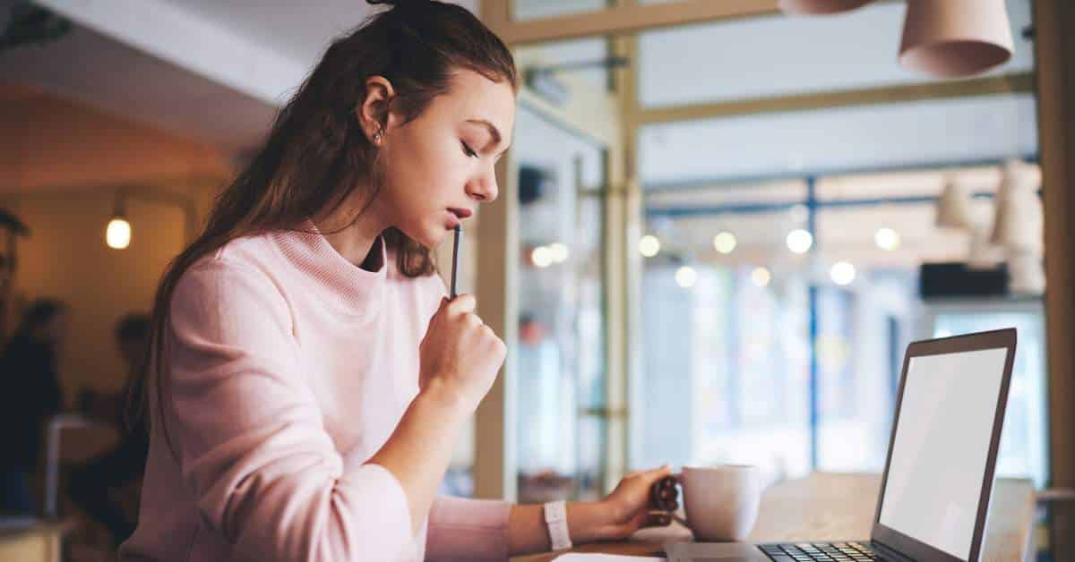 A teenage girl holds a pencil to her face and looks reflectively at her computer.