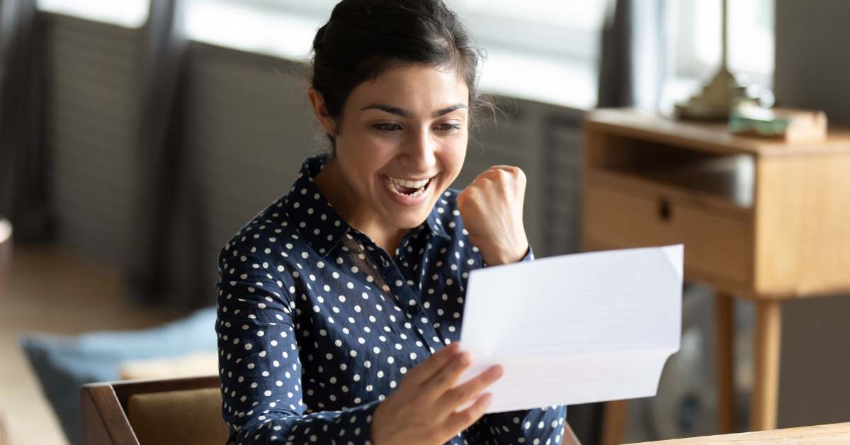 Young college-aged female in a navy blue shirt printed with white polka dots holds a paper in one hand and is drawing back her fist in success. Her mouth is open in happy surprise.