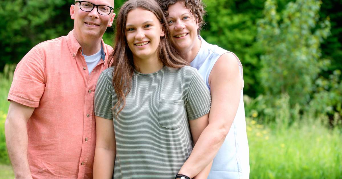 Balding father wearing glasses and a pink shirt, mother wearing a sleeveless shirt, each lean in to embrace a teenage daughter with long hair standing in the middle of them.