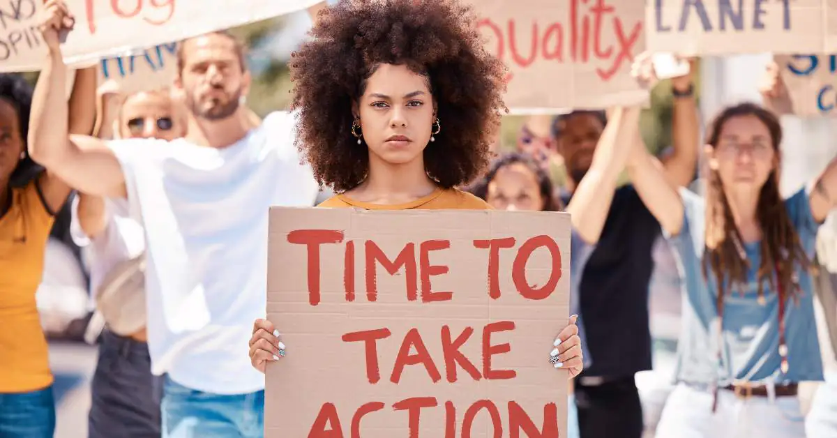 College students protesting and holding cardboard signs. The one in the forefront holds a sign that reads, "Time to Take Action"