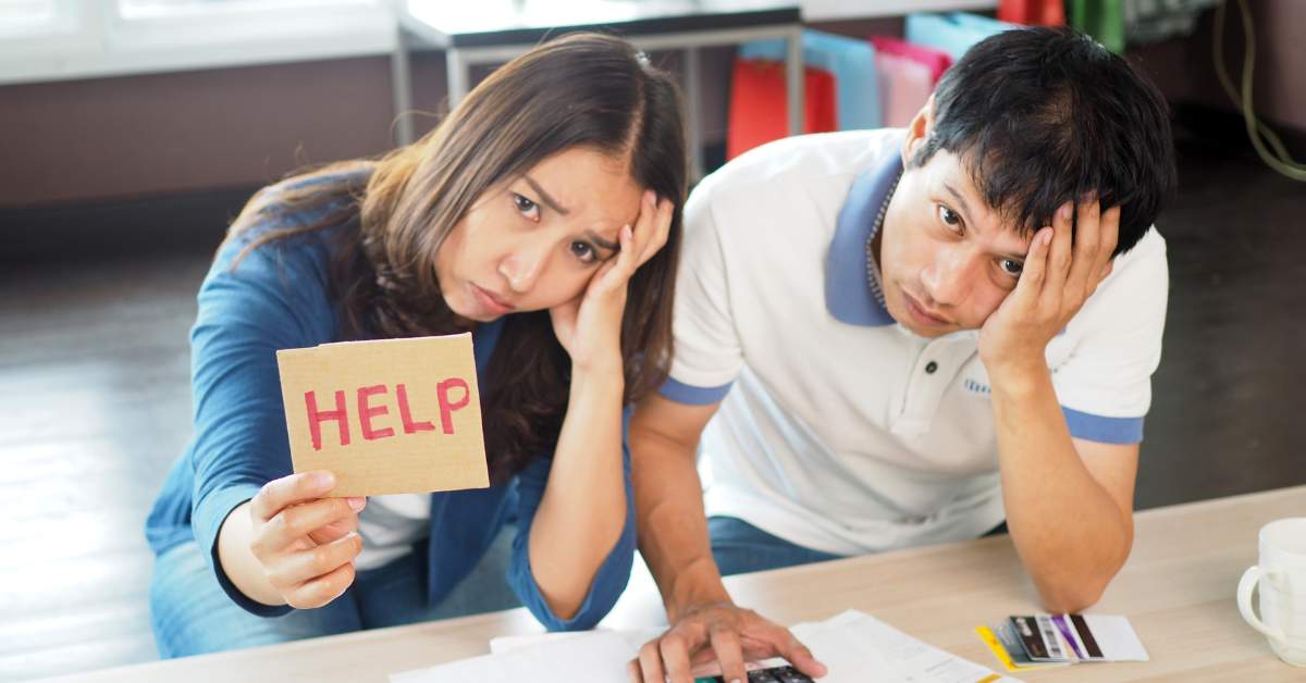 Middle aged man and woman, each with a hand on their forehead, holing up a cardboard sign that says "Help."