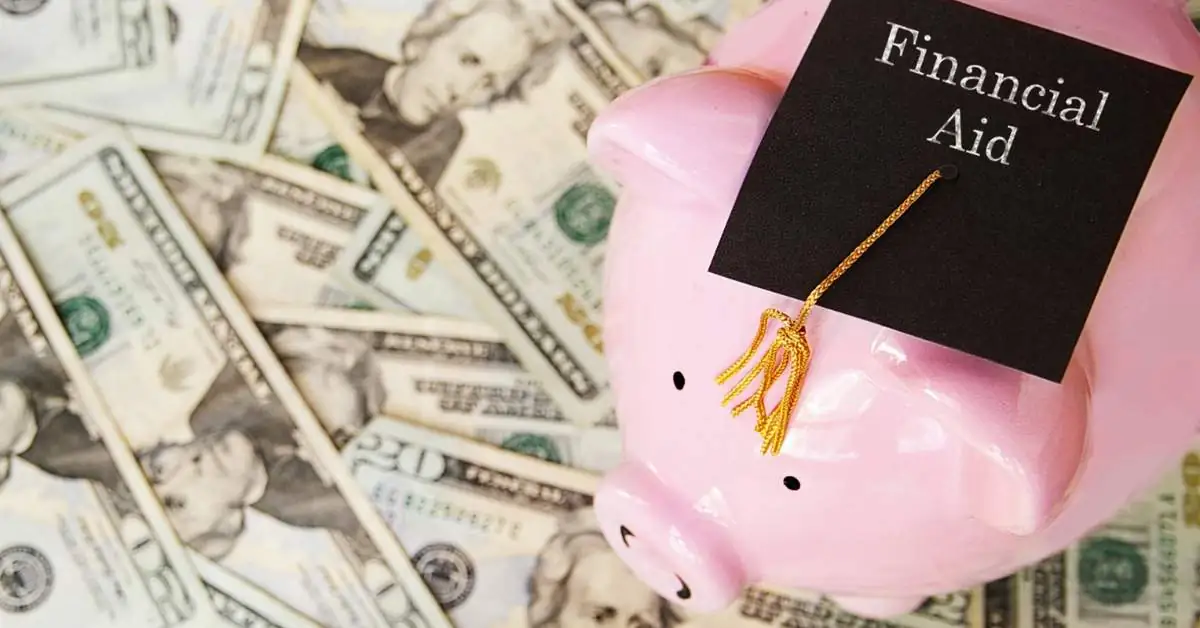 Money spread across a table with a pink piggy bank on top wearing a black mortar board that reads "Financial Aid."