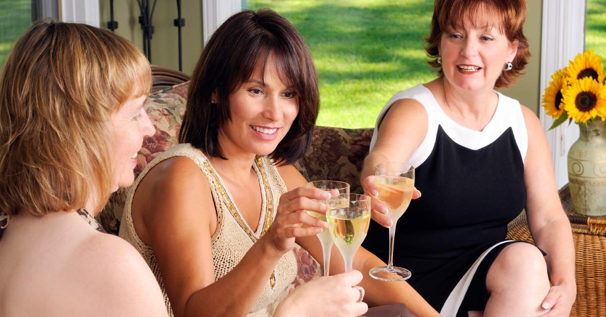 Three middle-aged women sitting on a couch, lifting their glass of alcohol in a toast