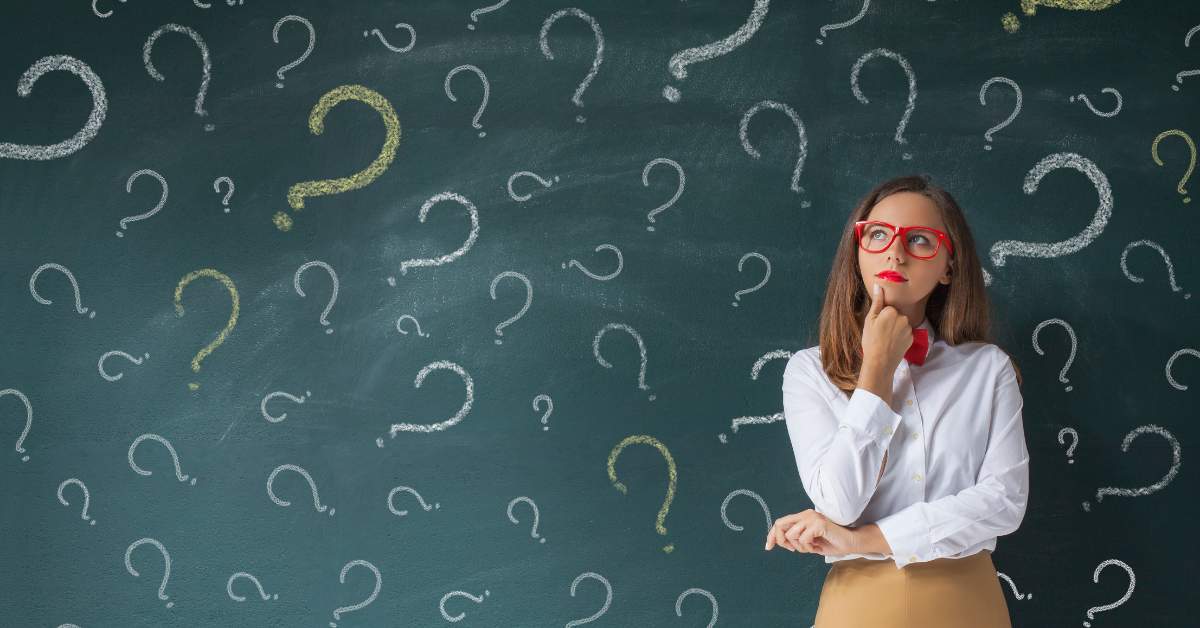 Professional woman with her fist on her chin looking up in contemplation. Behind her is a chalkboard filled with question marks.