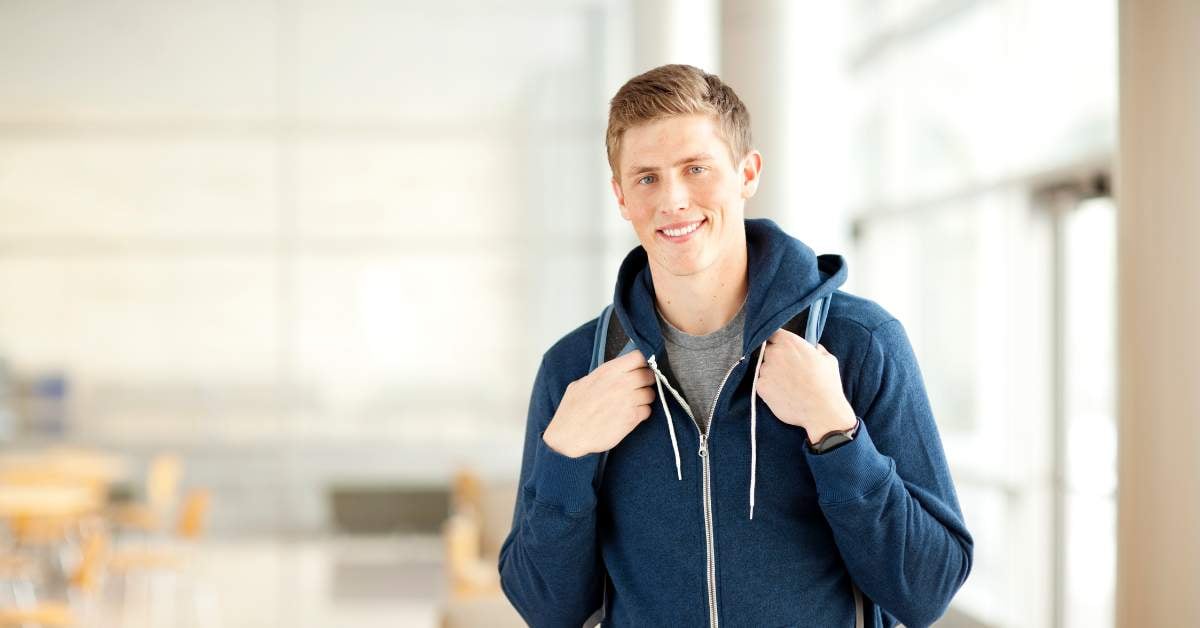 Blond young man standing in a classroom. He wears a blue zippered hoodie, has a backpack on, and is slightly smiling at the camera.