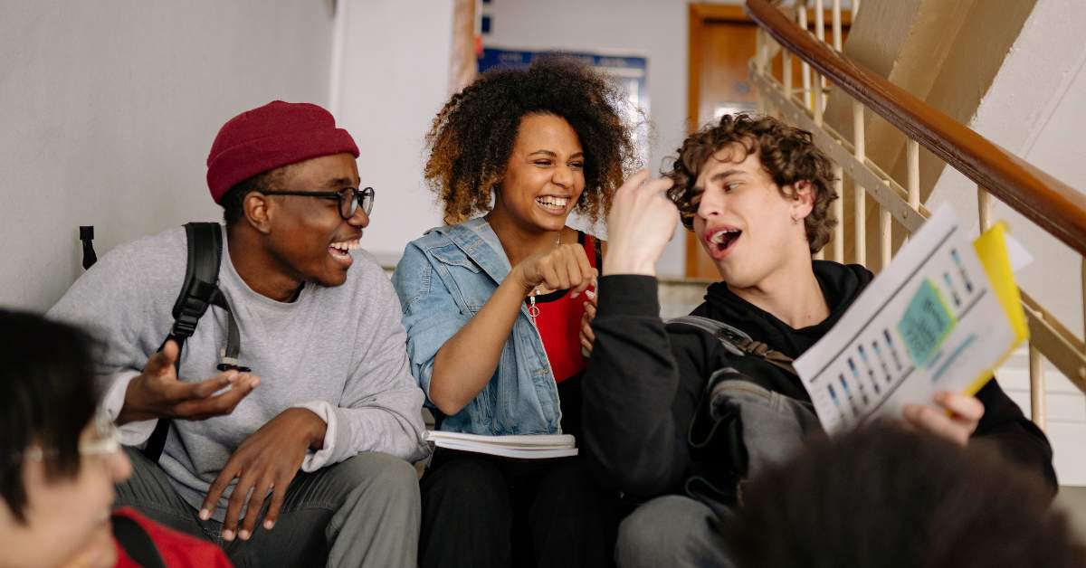 Three students, two men and one woman, sit on a coach laughing together.