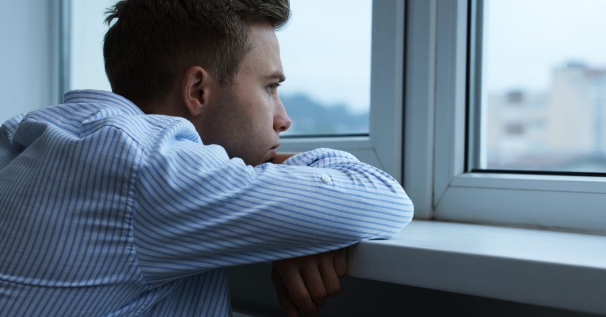 A young man wearing a blue striped button down shirt has his arms resting on a window ledge while he looks out. His chin is on his arms.